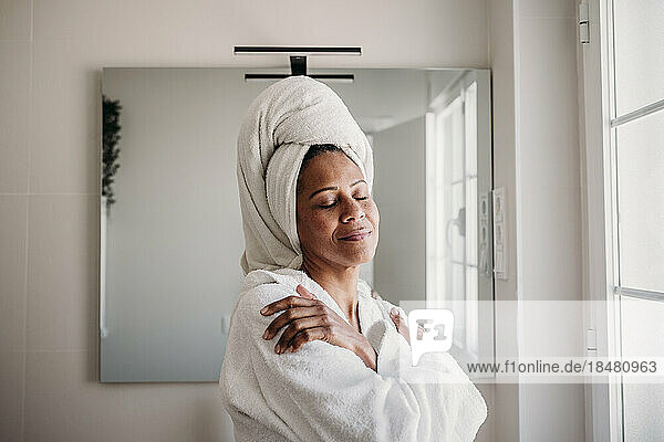 Smiling mature woman with eyes closed hugging self in bathroom at home