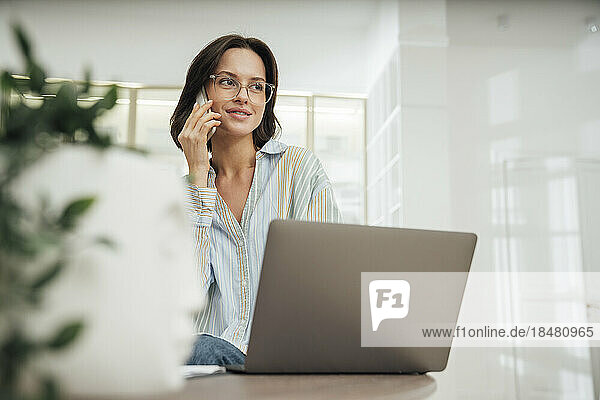 Smiling freelancer talking on smart phone and sitting with laptop at desk