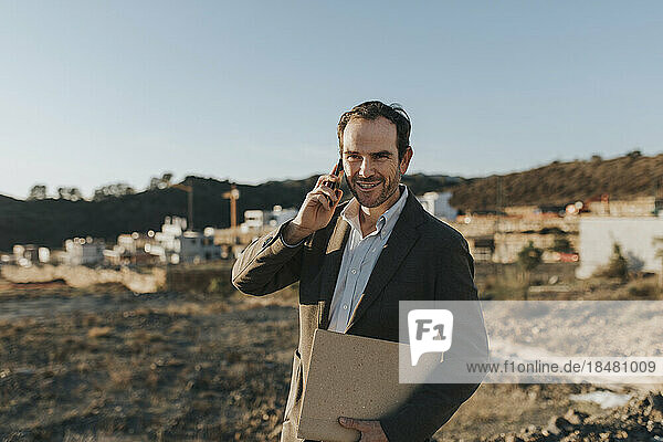 Smiling mature man talking over smart phone at empty area for residential construction