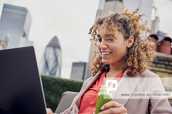 Happy young businesswoman looking at laptop holding smoothie