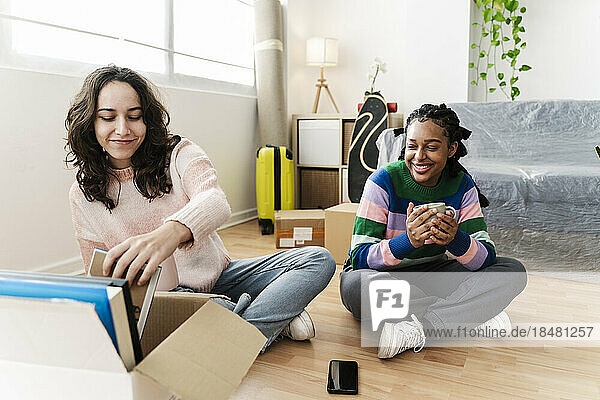 Smiling young woman unpacking box with friend holding coffee cup at home