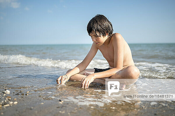 Boy playing in wet sand sitting cross-legged on shore at beach