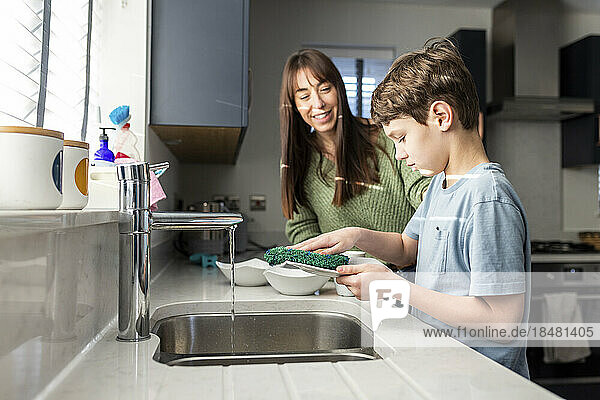 Happy woman with son washing dishes near kitchen sink