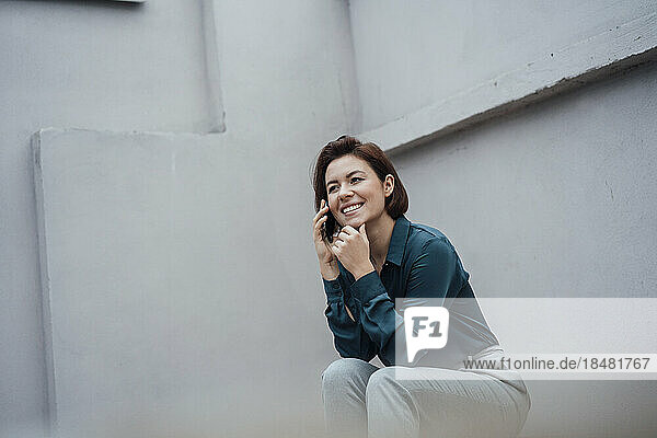 Smiling businesswoman talking over mobile phone in front of gray wall