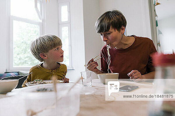 Son looking at mother eating noodles with chopsticks in home