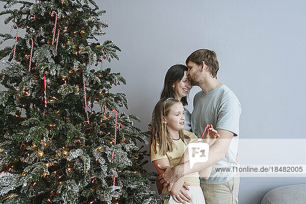 Mann küsst Frau und Tochter zu Hause am Weihnachtsbaum auf die Stirn