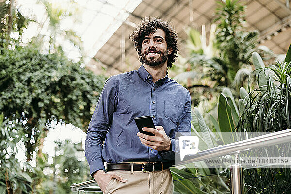 Smiling businessman with smart phone standing by railing in front of plants