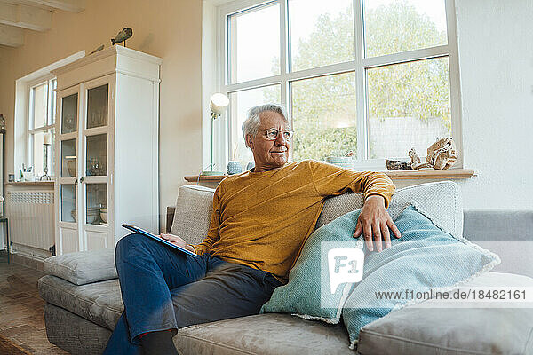 Senior man with tablet PC sitting on sofa in living room at home