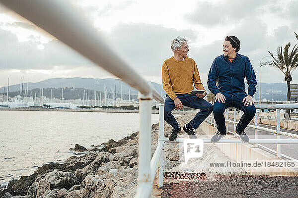 Father and son sitting on railing under cloudy sky