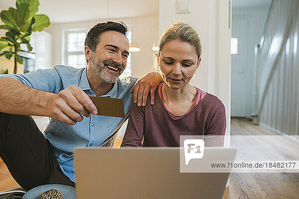 Happy husband holding credit card by woman using laptop at home