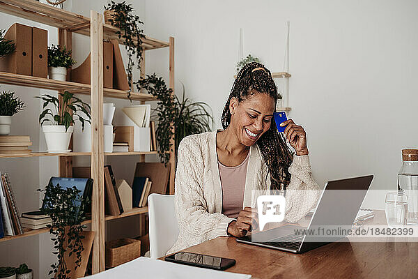 Happy freelancer sitting with credit card and laptop at desk