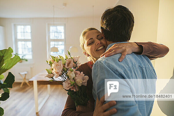 Smiling mature woman embracing man holding flowers at home