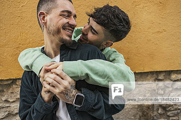 Affectionate gay man hugging boyfriend from behind in front of wall