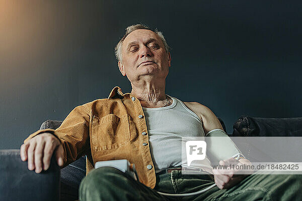 Senior man with eyes closed checking blood pressure sitting on sofa