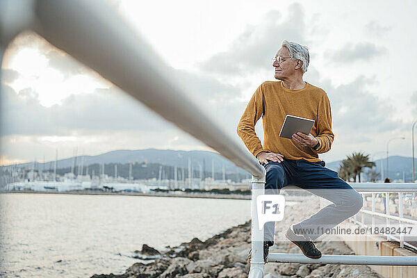 Smiling senior man sitting on railing with tablet PC