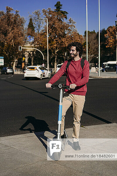 Glücklicher junger Mann steht mit Elektroroller auf der Straße