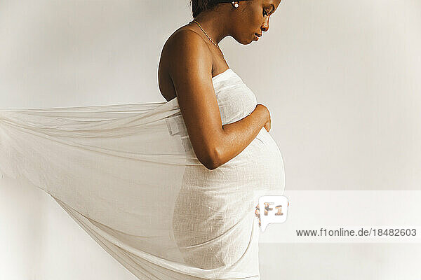 Young pregnant woman covered in white sheet in front of wall