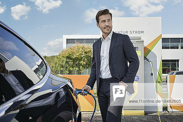 Smiling young businessman holding plug by car at electric vehicle charging station
