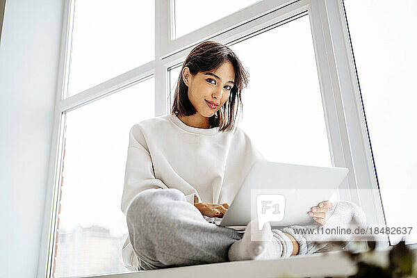 Smiling young woman sitting with laptop in front of window at home