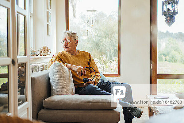 Nachdenklicher älterer Mann mit kabellosen Kopfhörern sitzt zu Hause auf dem Sofa