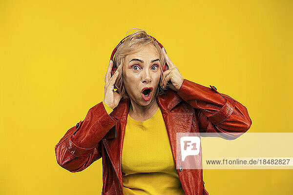 Shocked senior woman wearing wireless headphones listening to music against yellow background