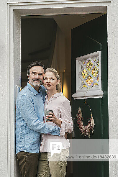 Mature man embracing wife holding cup at doorway of house