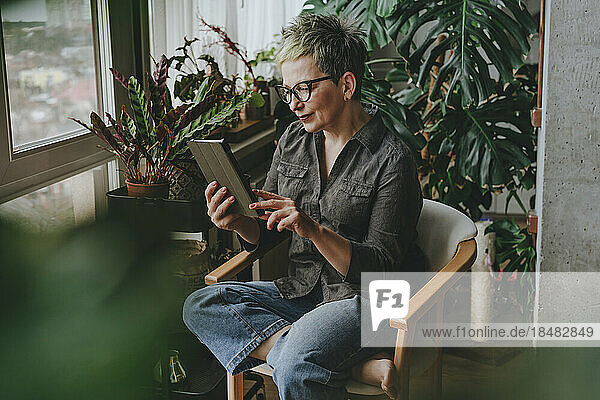 Reife Frau sitzt zu Hause mit Tablet-PC auf einem Stuhl vor Pflanzen