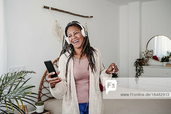 Glückliche Frau mit kabellosen Kopfhörern steht im Schlafzimmer