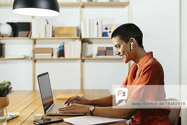 Smiling freelancer wearing in-ear headphones working on laptop at table