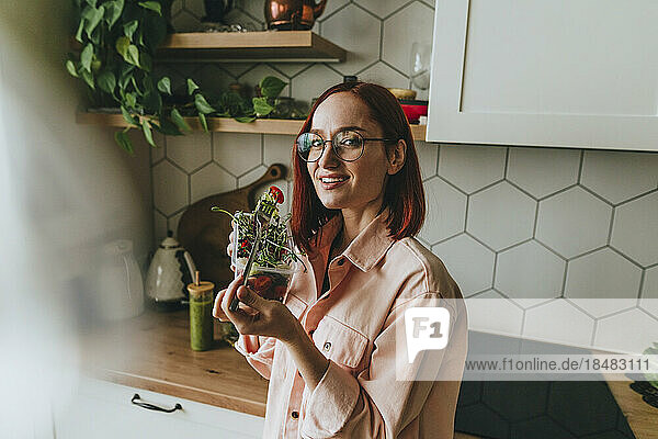 Glückliche Frau mit Brille  die zu Hause in der Küche Salat isst