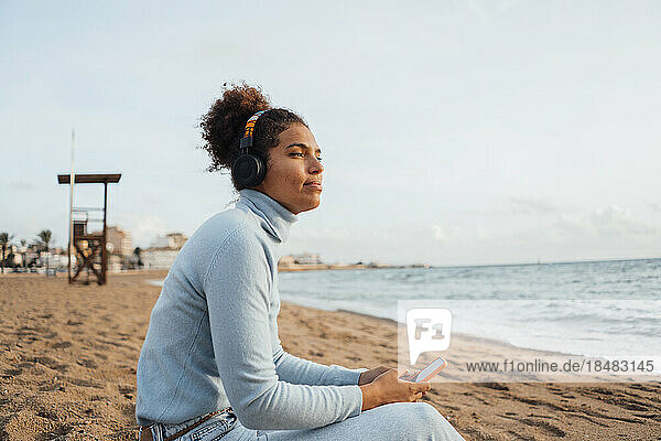 Nachdenkliche junge Frau sitzt mit Smartphone am Strand