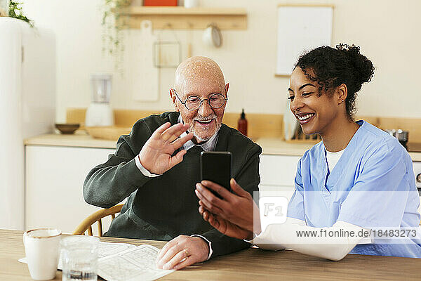 Caretaker holding smart phone for senior man waving on video call at home