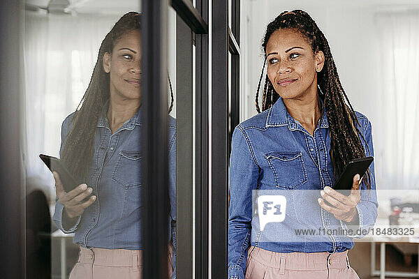 Thoughtful mature woman standing with smart phone by glass door at home