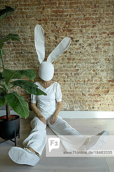 Frau mit Kaninchenmaske sitzt neben Pflanze und Ziegelmauer