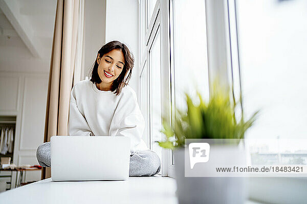 Smiling young woman using laptop on window sill