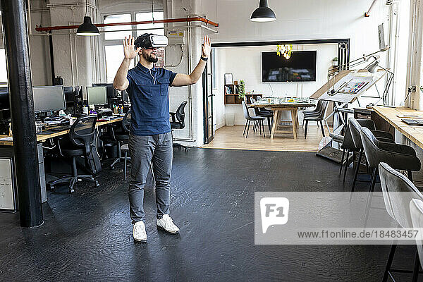 Young businessman wearing virtual reality simulator standing with hands raised in office