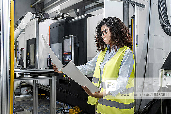 Young engineer examining blueprints standing by machine in industry