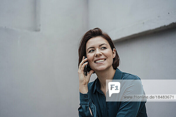 Happy businesswoman talking through smart phone by gray wall