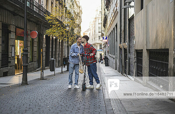 Glückliches schwules Paar küsst sich mit einem elektrischen Tretroller inmitten von Gebäuden