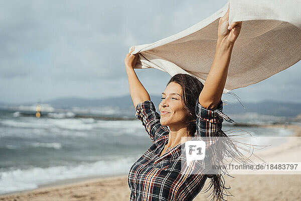 Lächelnde Frau mit Schal und erhobenen Armen am Strand