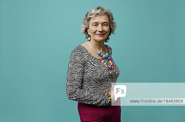 Smiling senior woman wearing multi colored jewelry against turquoise background