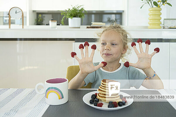 Girl with raspberries on fingers sitting at dining table in kitchen