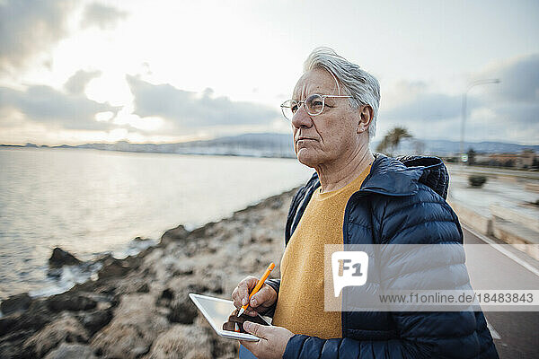 Senior man standing with tablet PC in front of sea at promenade