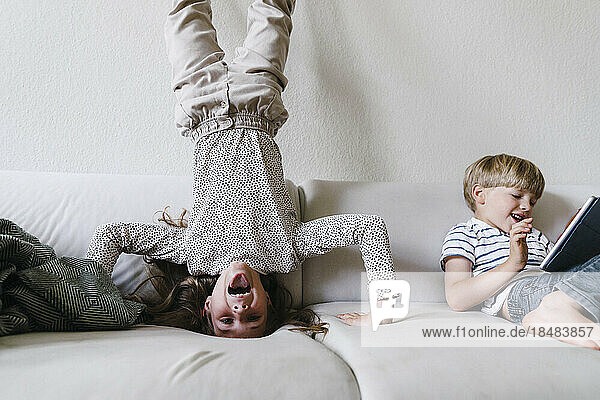 Carefree girl practicing headstand by brother on sofa at home