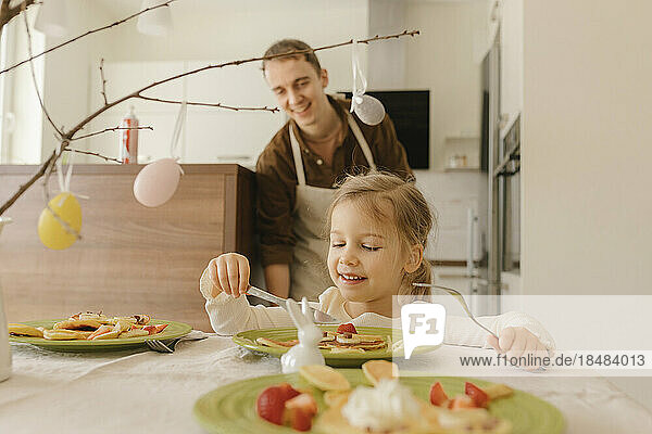Smiling girl eating pancakes on table at home