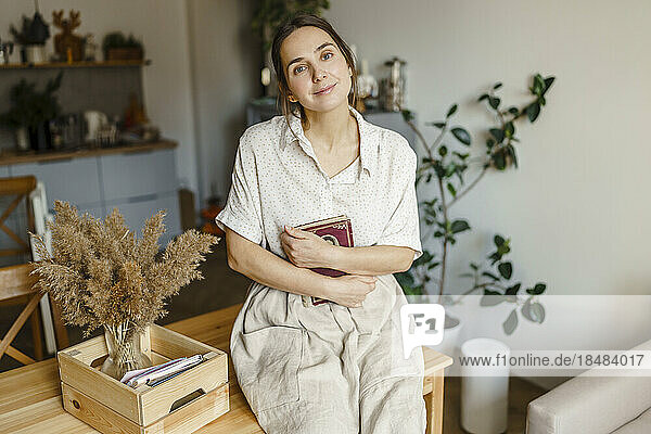 Lächelnde Frau mit Buch neben Vase auf dem Tisch im Wohnzimmer zu Hause