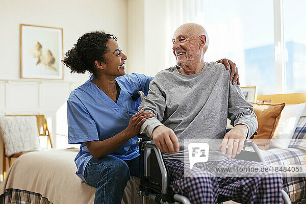 Happy nurse with senior man sitting on wheelchair in bedroom at home