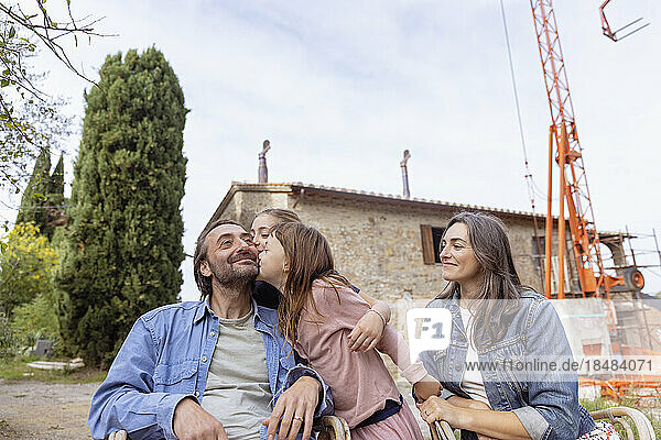 Girls kissing father with mother sitting in front of house