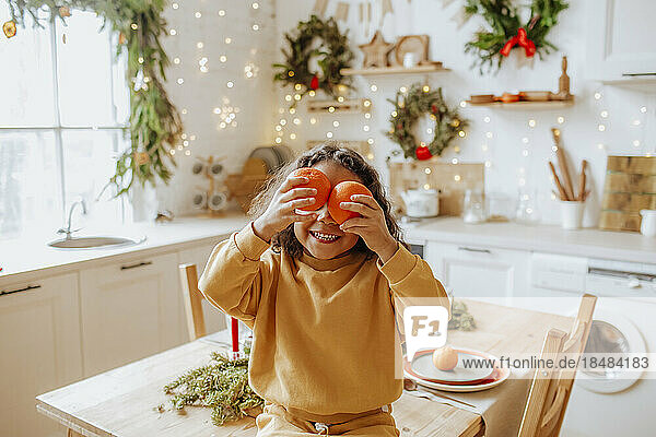 Girl holding oranges over eyes sitting on table at home
