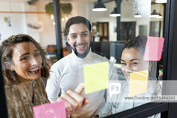Smiling businesswoman with colleagues reading adhesive notes on glass in office